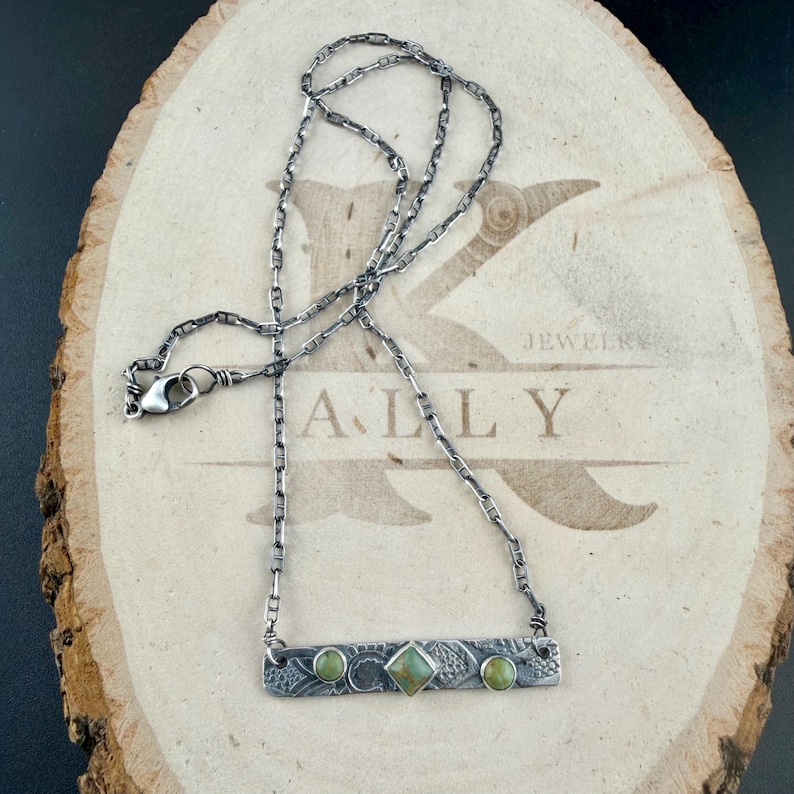 May include: A silver-toned necklace featuring a rectangular bar pendant with an oxidized finish, textured patterns, and three inlaid green stones. The center stone is square-shaped, flanked by two round stones. The chain is a linked cable style.