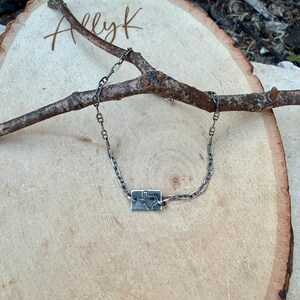 May include: A silver chain bracelet with a rectangular pendant featuring a cutout of the state of Texas. The bracelet is displayed on a wooden surface with a tree branch.
