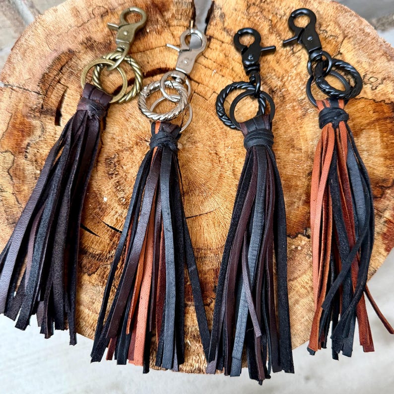 May include: Four leather tassel keychains in shades of brown and black. Each keychain has a different metal clasp and ring. The tassels are made of strips of leather, and the keychains are arranged on a wooden surface.