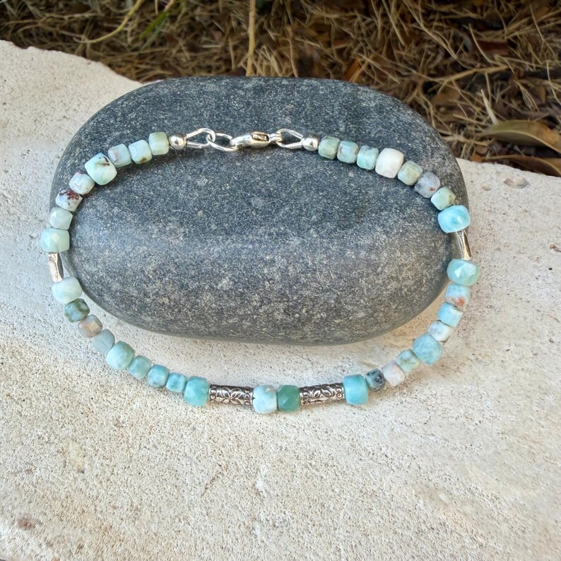 Puede incluir: Una pulsera de cuentas con peque&ntilde;as piedras cuadradas de color azul claro y verde azulado, intercaladas con dos cuentas cil&iacute;ndricas de metal plateado grabado. La pulsera se asegura con un cierre de mosquet&oacute;n de metal plateado.