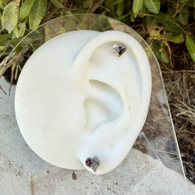 May include: Two small silver-toned heart-shaped stud earrings displayed on a white ear-shaped model. One earring is positioned in the upper cartilage area, and the second earring is placed in the lower earlobe position on the white model.