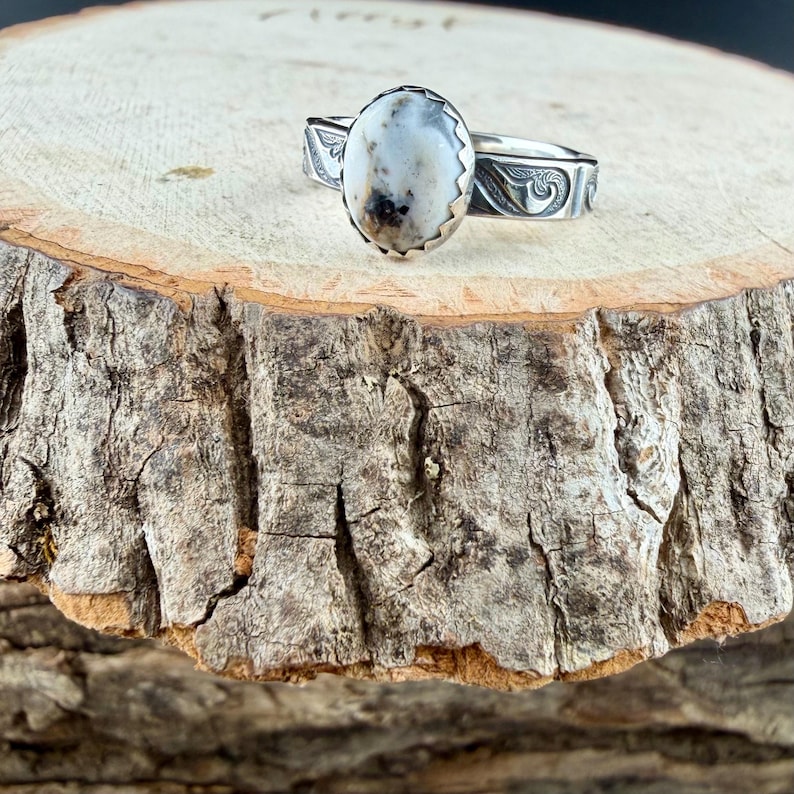 May include: A silver ring with an oval, white and brown stone. The ring has a decorative band with etched designs. The ring is displayed on a wooden surface.