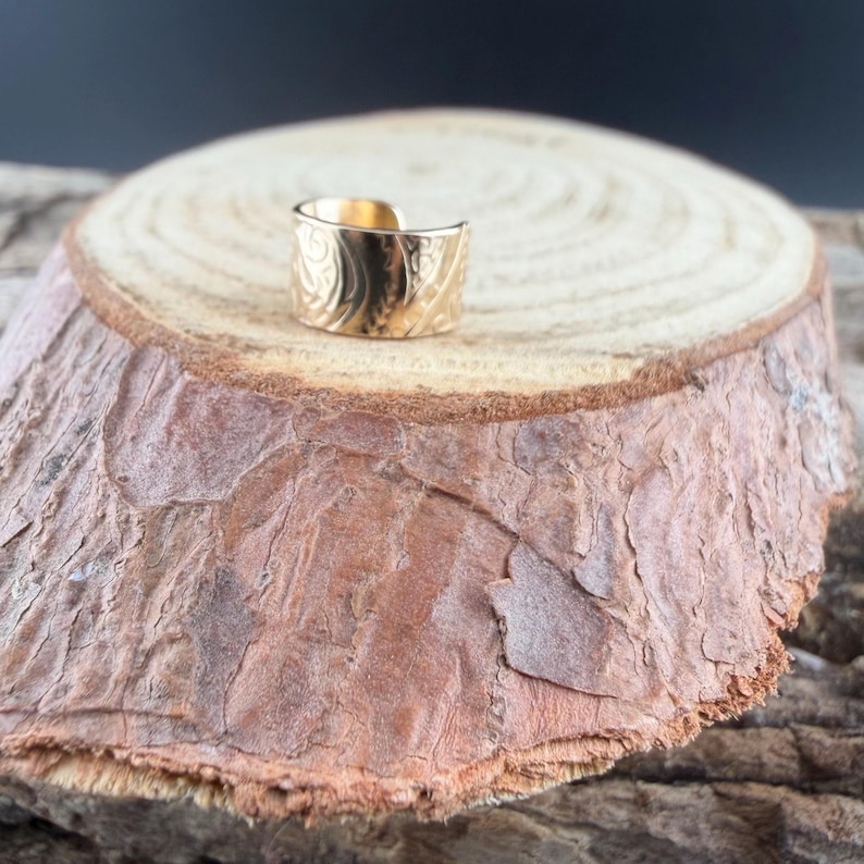 May include: A gold-colored ring with an engraved floral pattern. The ring is displayed on a wooden surface with a natural, rustic appearance. The ring's design features intricate details, and the open-ended style allows for adjustable sizing.