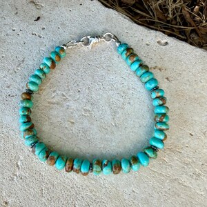 May include: A beaded bracelet featuring round, turquoise-colored stones with brown matrix inclusions. The bracelet is strung on a cord and secured with a silver-toned metal lobster claw clasp, showcasing a consistent pattern of natural-looking beads.