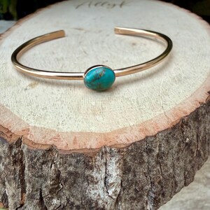 May include: A gold-colored cuff bracelet with a turquoise stone set in the center. The bracelet is open-ended and rests on a wooden surface. The turquoise stone is oval-shaped and has a mottled appearance.
