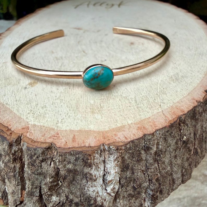 May include: A gold-colored cuff bracelet with a turquoise stone set in the center. The bracelet is open-ended and rests on a wooden surface. The turquoise stone is oval-shaped and has a mottled appearance.