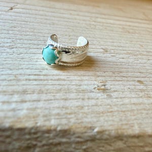 May include: A silver open-band ring featuring a round, polished turquoise stone set in a scalloped bezel. The band has a textured, grooved edge and a smooth, polished finish, with a gap at the back allowing for adjustable sizing.