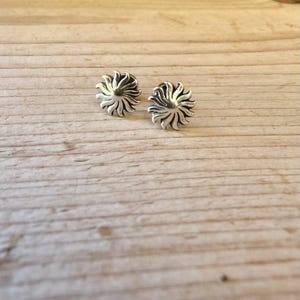 May include: A pair of silver stud earrings shaped like stylized suns with radiating curved rays. The metal has a polished finish with darkened recessed areas between the rays to create depth and contrast in the circular sun design.