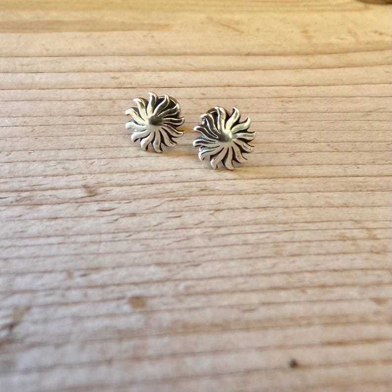 May include: A pair of silver stud earrings shaped like stylized suns with radiating curved rays. The metal has a polished finish with darkened recessed areas between the rays to create depth and contrast in the circular sun design.