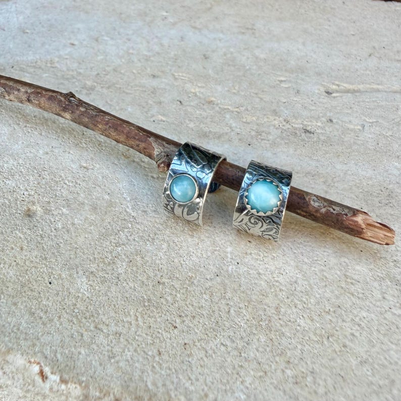 May include: A pair of silver hoop earrings featuring a wide, textured band with an intricate, dark, etched pattern. Each earring is centered with a smooth, round, light blue cabochon stone set in a scalloped bezel, resting on a wooden branch.