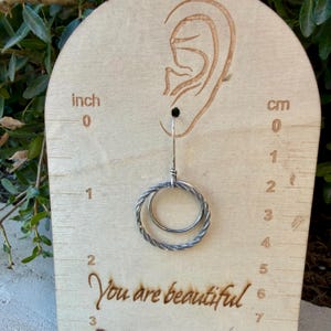 May include: A silver-toned earring featuring two interlocking circular hoops, one with a smooth finish and the other with a twisted texture, displayed on a wooden card with an ear outline and a ruler showing measurements in inches and centimeters.