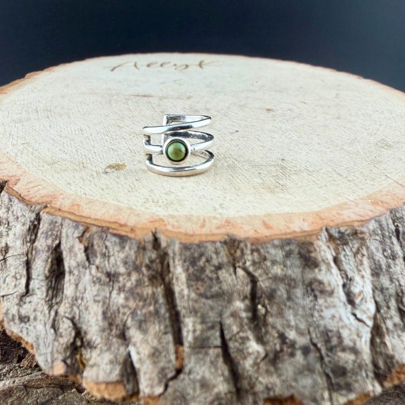 May include: A silver-colored ear cuff with a spiral design, featuring a small, round, olive-green stone at the center. The jewelry is displayed on a rustic, wooden surface with a natural bark edge.