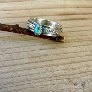 Puede incluir: Un anillo de plata con una piedra turquesa ovalada central con vetas de matriz marr&oacute;n. La banda est&aacute; hecha de plata pulida y muestra un patr&oacute;n en relieve de peque&ntilde;as flores y l&iacute;neas verticales que rodean toda la superficie exterior.