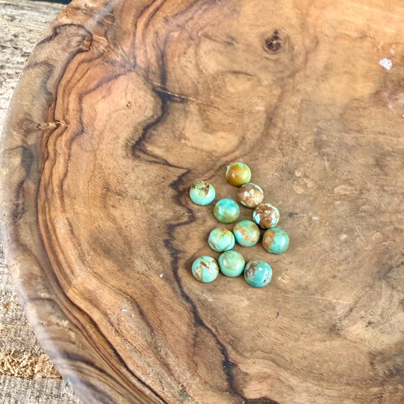 May include: A selection of small, round turquoise gemstones with brown and tan markings. The stones are arranged on a wooden bowl with a natural wood grain. The gemstones range in colour from light to dark turquoise.