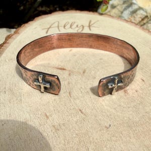 May include: A copper cuff bracelet featuring a hammered texture and a polished finish. The open-ended design displays two small, silver-toned cross emblems positioned at each terminal end of the band, creating a distinct contrast in metal color.