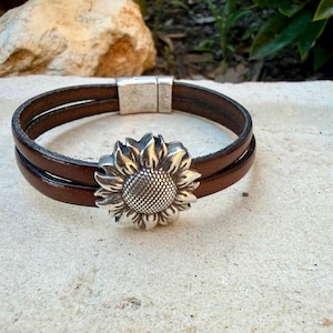 May include: Brown leather bracelet with a silver sunflower charm. The bracelet features two parallel leather bands and a magnetic clasp. The sunflower charm is detailed with textured petals and a central seed pattern. A stylish accessory.