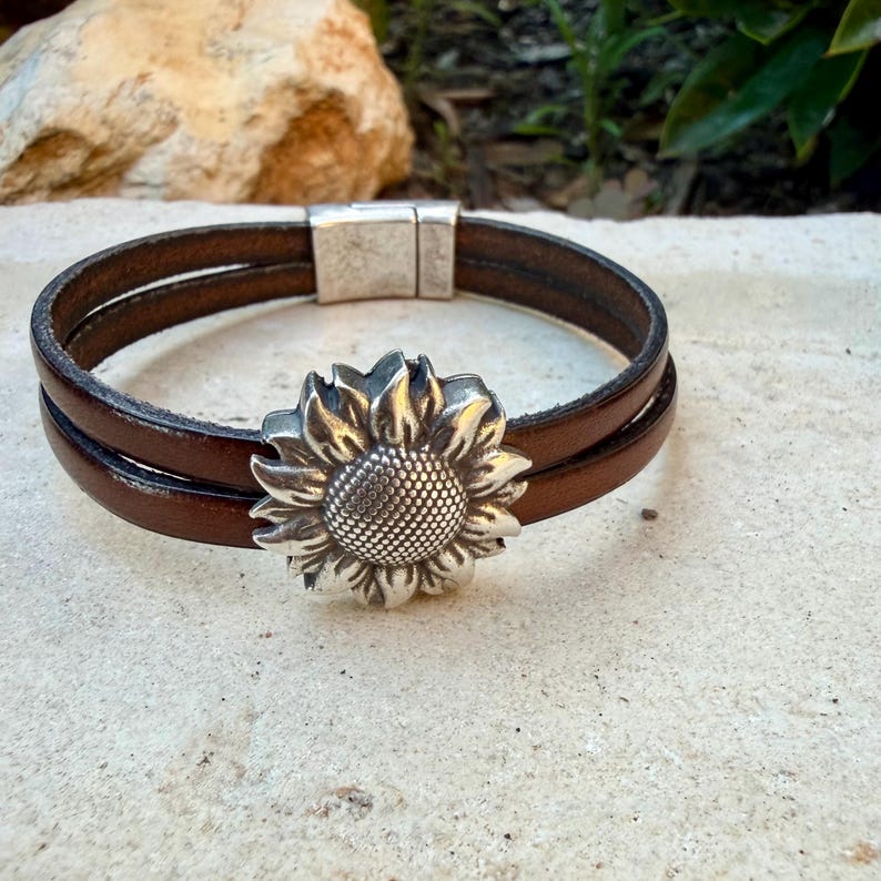 May include: Brown leather bracelet with a silver sunflower charm. The bracelet features two parallel leather bands and a magnetic clasp. The sunflower charm is detailed with textured petals and a central seed pattern. A stylish accessory.