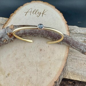 May include: Gold-toned cuff bracelet with a small, round, gray gemstone. The open-ended bracelet is displayed on a wooden surface with the handwritten text "Allyk". The jewelry is simple and elegant.