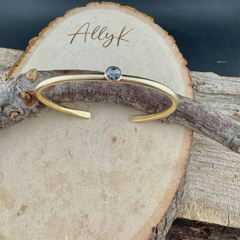 May include: Gold-toned cuff bracelet with a small, round, gray gemstone. The open-ended bracelet is displayed on a wooden surface with the handwritten text "Allyk". The jewelry is simple and elegant.