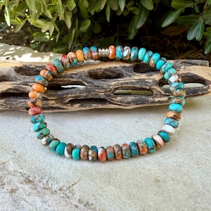 May include: A beaded bracelet featuring small, round, flattened stones in shades of turquoise, orange, white, and brown. The beads are arranged in a circular pattern and include a single metallic silver-toned spacer bead near the top center.