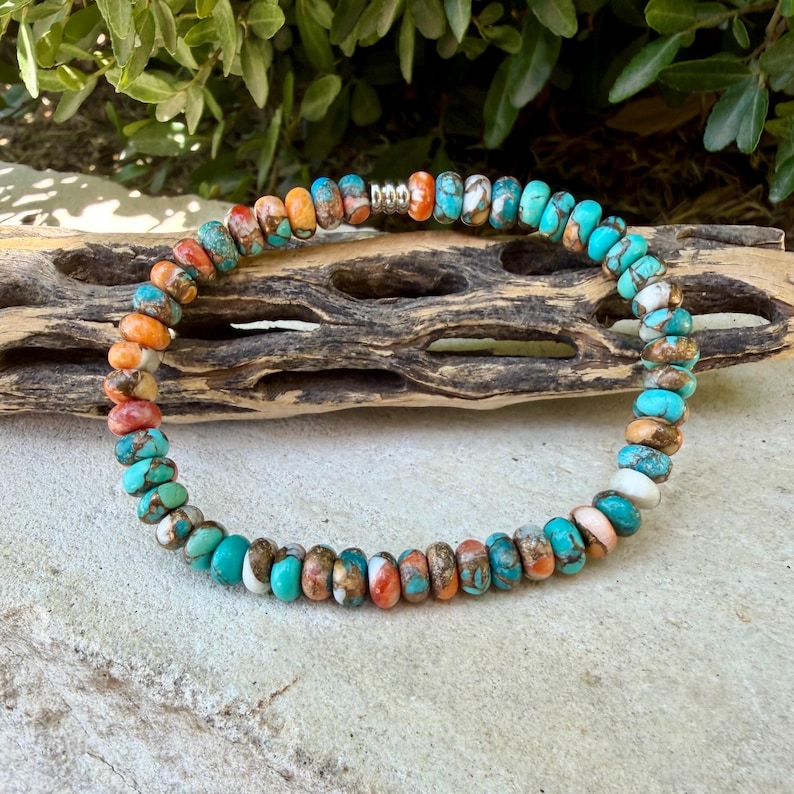 May include: A beaded bracelet featuring small, round, flattened stones in shades of turquoise, orange, white, and brown. The beads are arranged in a circular pattern and include a single metallic silver-toned spacer bead near the top center.