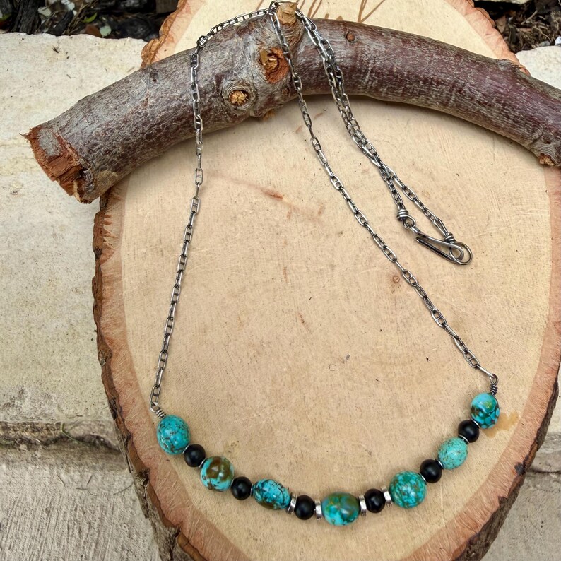 May include: A silver chain necklace with turquoise and black beads. The necklace is displayed on a wooden surface with a piece of wood in the background. The turquoise beads are a vibrant blue-green color, and the black beads are interspersed throughout the design.
