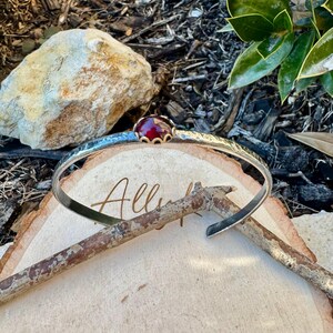 Puede incluir: Un brazalete plateado con textura martillada y una gema roja engastada en un bisel dorado. El brazalete abierto descansa sobre una superficie de madera con la palabra "All" visible. Di&aacute;metro aproximado de 15 cm.