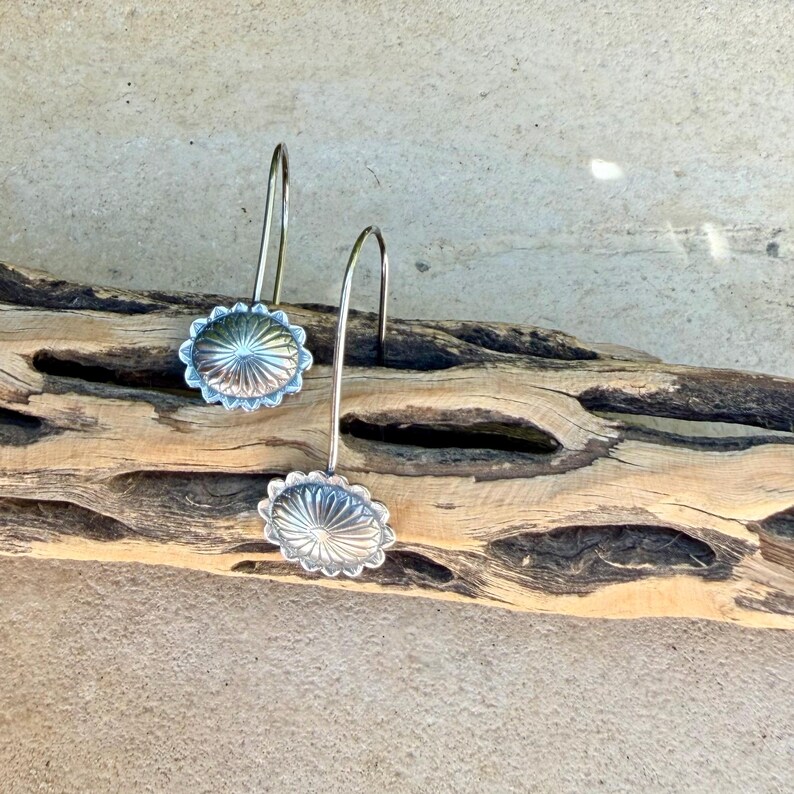 May include: A pair of silver-toned drop earrings featuring circular, sunburst-patterned medallions with scalloped edges. Each earring is suspended from a curved wire hook, showcasing a polished metallic finish with intricate radial line detailing.