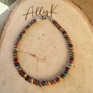 May include: A beaded bracelet with a variety of earth-toned beads, including red, green, yellow, and brown. The bracelet is strung on a silver chain and has a silver clasp. The word "Allyk" is written in cursive on the wooden surface.