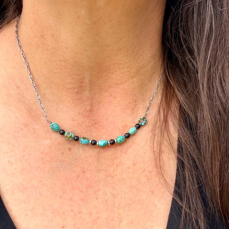 May include: A silver chain necklace featuring turquoise and black beads. The necklace has a delicate chain and is adorned with a series of round, turquoise-colored beads interspersed with black and silver beads. The necklace is worn around the neck.