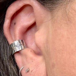 Puede incluir: Primer plano de una oreja adornada con un brazalete de plata y un peque&ntilde;o pendiente de bot&oacute;n. El brazalete es ancho, con un dise&ntilde;o geom&eacute;trico. El pendiente de bot&oacute;n tiene forma cuadrada. El tono de piel es claro.