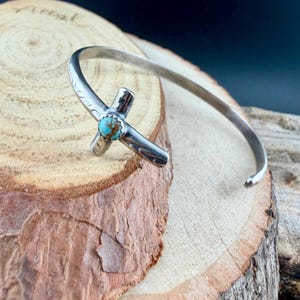 May include: A silver cuff bracelet with a cross-shaped centerpiece. The cross features a turquoise stone at its center. The bracelet is resting on a wooden surface, showcasing its handcrafted design and Southwestern style.