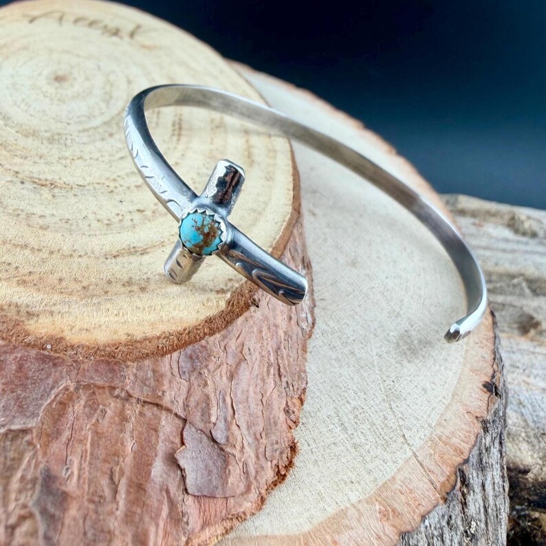 May include: A silver cuff bracelet with a cross-shaped centerpiece. The cross features a turquoise stone at its center. The bracelet is resting on a wooden surface, showcasing its handcrafted design and Southwestern style.