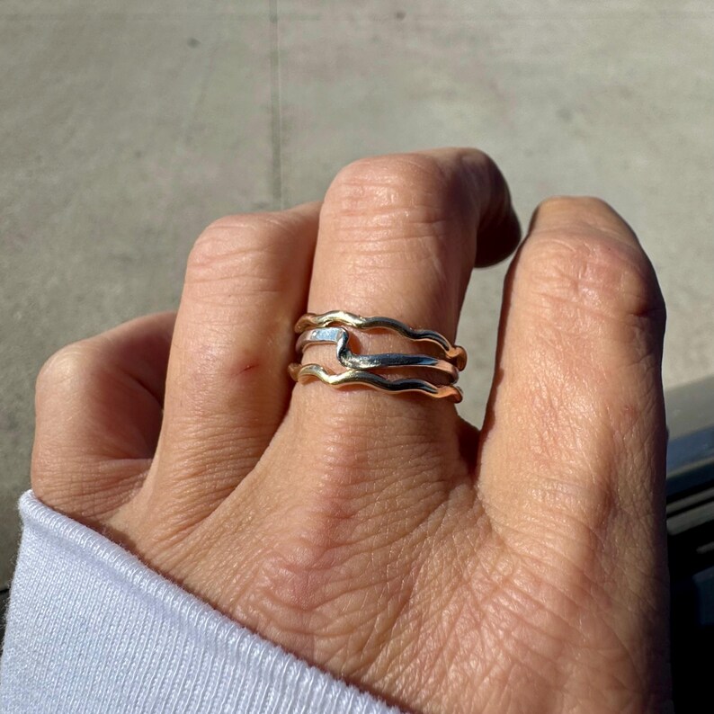 Puede incluir: Un conjunto de tres anillos con un dise&ntilde;o ondulado. Los anillos superior e inferior son de color dorado, mientras que el anillo central es de color plateado. Los anillos est&aacute;n apilados en un dedo, mostrando su forma &uacute;nica y fluida. Joyas.
