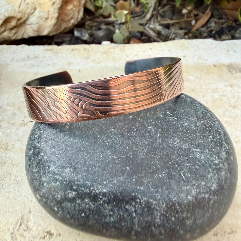 May include: A copper cuff bracelet with an etched wave pattern. The bracelet is displayed on a dark gray stone. The metal has a warm, reddish-brown hue, and the etched design adds texture and visual interest to the jewelry.