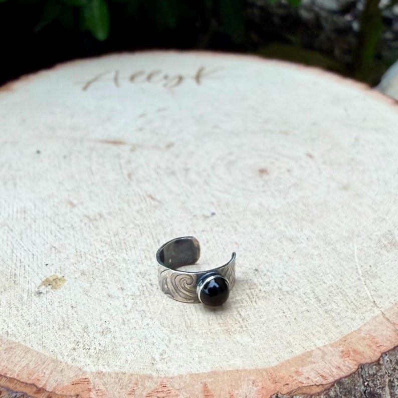 May include: A silver-toned adjustable ring with a black gemstone. The ring features a swirling pattern on the band and a round, polished black stone. The ring is displayed on a wooden surface.