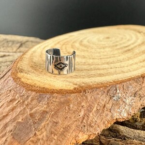 May include: A silver-colored ring with a textured surface and an engraved geometric design. The ring is open-ended and sits on a wooden surface, showcasing its details and craftsmanship. The ring's design is a central focus.