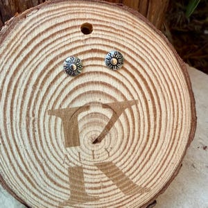 May include: A pair of small, round silver stud earrings featuring a raised sun design with radiating rays. The earrings are displayed on a circular wooden slice with visible growth rings and the engraved text "ALLY" visible below them.