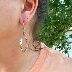 Op de afbeelding: Een zilverkleurige hangende oorbel met een cirkelvormig ringontwerp met een gevlochten of gedraaide metaalstructuur. De oorbel hangt aan een eenvoudige haak en heeft een gepolijste metalen afwerking die het licht op subtiele wijze vangt.