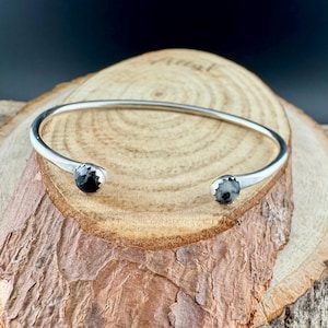 May include: A silver cuff bracelet with a simple design. The open bracelet has a smooth, curved band with a small, dark, textured stone at each end. The bracelet is displayed on a piece of wood.