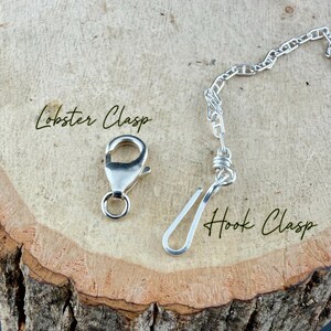 May include: Close-up of two types of jewelry clasps: a silver lobster clasp and a hook clasp. The clasps are on a wooden surface. The words "Lobster Clasp" and "Hook Clasp" are written in cursive.