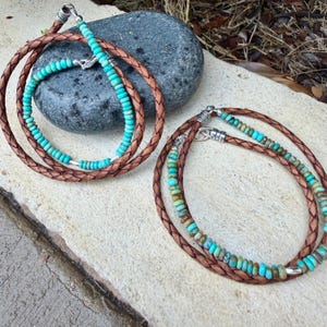May include: Two braided brown leather cords featuring turquoise heishi beads and silver-toned metal accents. Each cord is arranged in a circular loop, showcasing a combination of natural brown leather texture and vibrant blue-green stone beads.