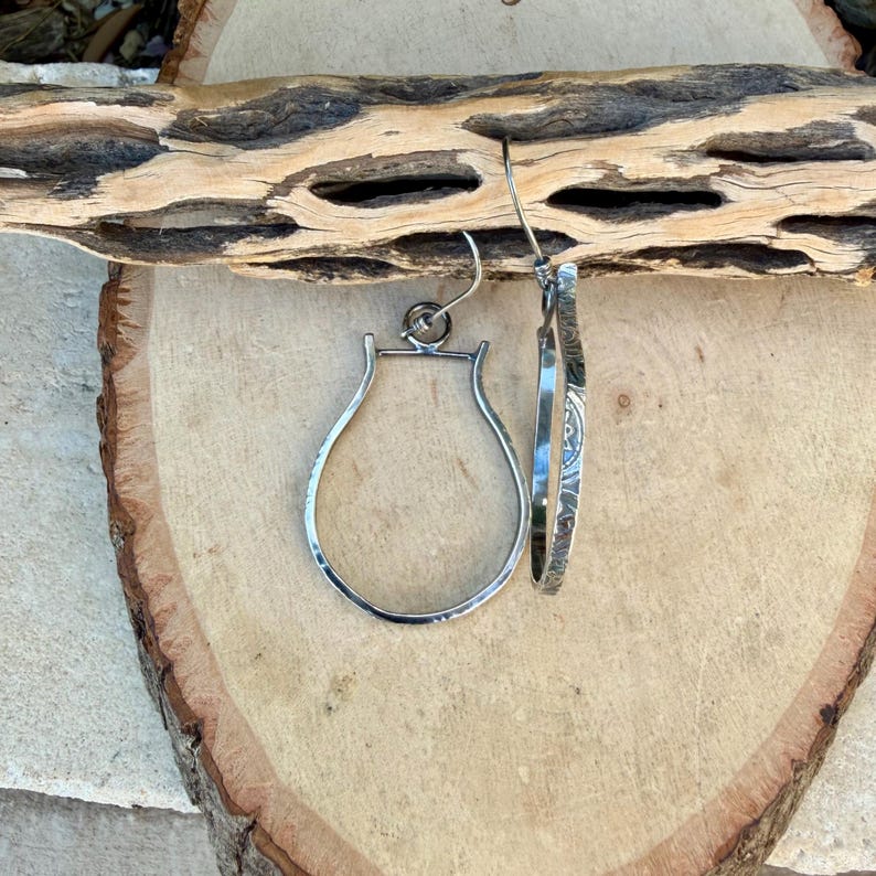 May include: A pair of silver hoop earrings with a unique, open teardrop design. One earring features an etched pattern. The earrings are displayed on a wooden surface with a natural, rustic aesthetic.