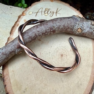 May include: A copper-colored cuff bracelet with a twisted design. The bracelet is resting on a wooden surface with the handwritten text "Allyk". The bracelet's design features two strands of copper that are intertwined.