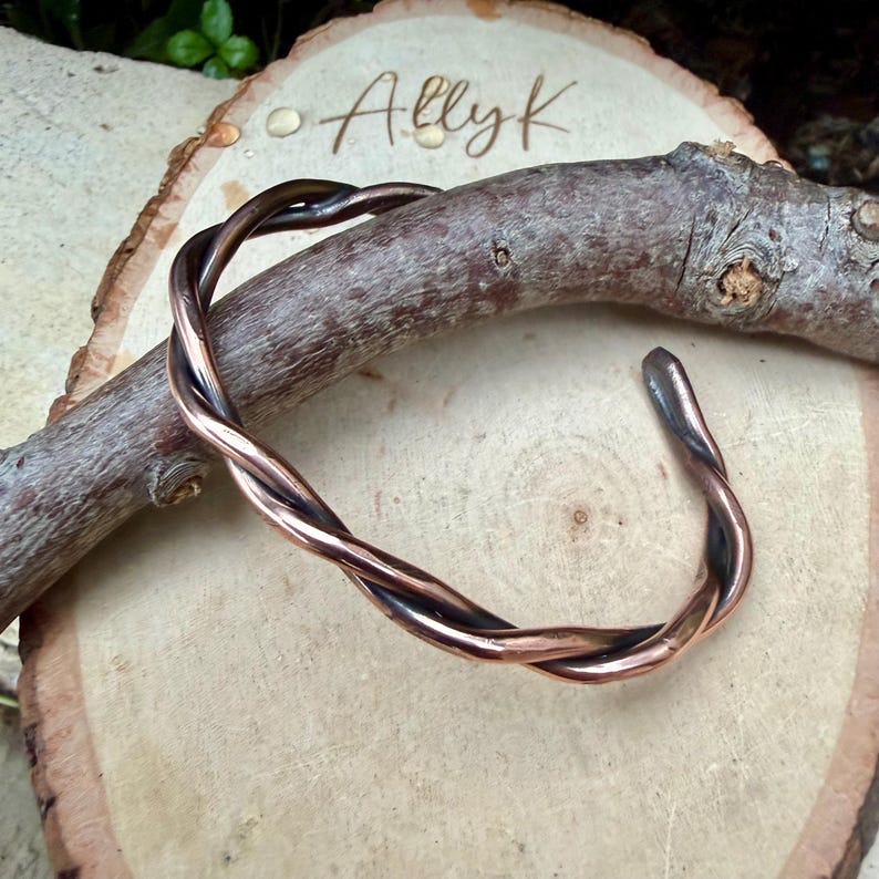 May include: A copper-colored cuff bracelet with a twisted design. The bracelet is resting on a wooden surface with the handwritten text "Allyk". The bracelet's design features two strands of copper that are intertwined.