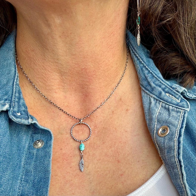 May include: Silver-toned necklace with a circular pendant, turquoise bead, and feather charm. The necklace is worn over a denim jacket. A matching earring with a feather charm is also visible.