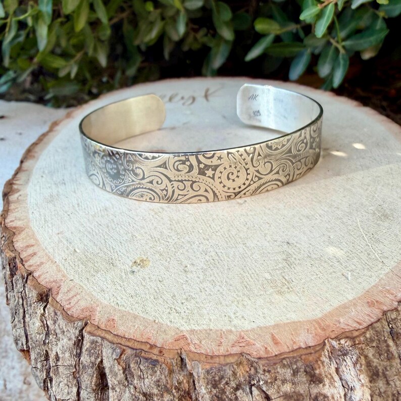 May include: A silver cuff bracelet featuring an intricate, engraved paisley and swirl pattern across its outer surface. The interior of the metal band is stamped with the letters AK and the number 925, indicating its sterling silver composition.
