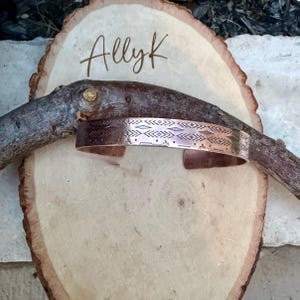 May include: A copper cuff bracelet featuring a repeating geometric pattern of diamonds and chevrons. The bracelet is positioned over a wooden branch resting on a light-colored wood slice that has the word AllyK engraved on its surface.