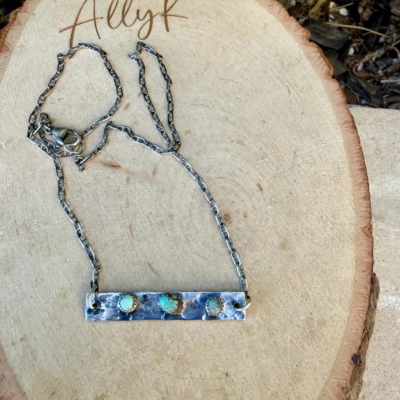 May include: A silver-toned necklace with a rectangular pendant featuring three turquoise stones. The chain is a linked design, and the pendant has a hammered texture. The word "Allyk" is written in cursive at the top.