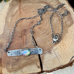 May include: A silver necklace featuring a rectangular metal bar pendant with an embossed floral pattern and a central oval iridescent cabochon stone. The pendant is attached to a silver chain with a lobster claw clasp closure.